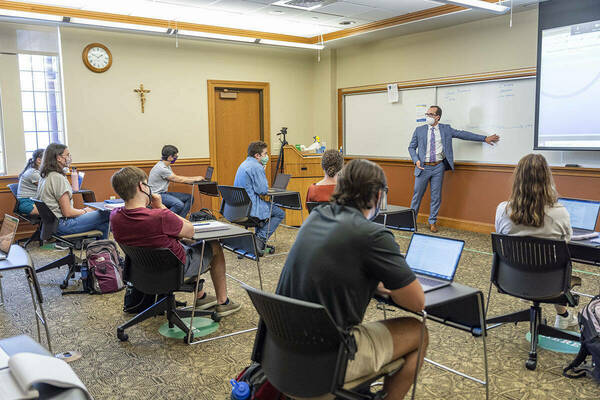Students seated at individual desks face away from the camera, looking toward speaker at the front of a classroom