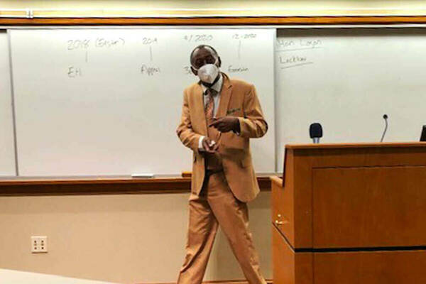 An individual wearing a light brown suit stands at the front of a classroom, addressing the audience