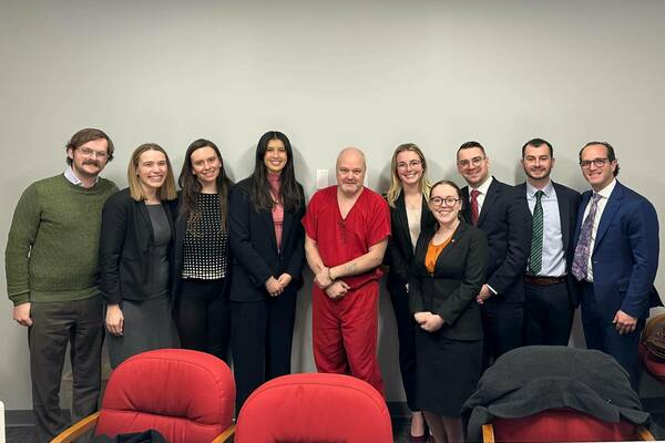 Ten individuals, including one wearing red prison attire, stand together for a group photo in a room with a plain wall and red chairs.