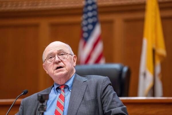 Older bald man in glasses, blue shirt, gray pinstripe suit, red striped tie, speaking at a microphone. American flag in background.